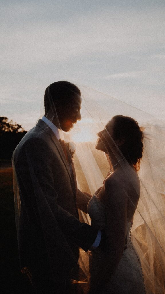 A bride and groom hold each other with a veil over both heads, outside at sunset.