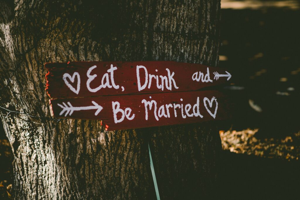 A sign nailed to a tree at a wedding, it says "Eat Drink & Be Married"