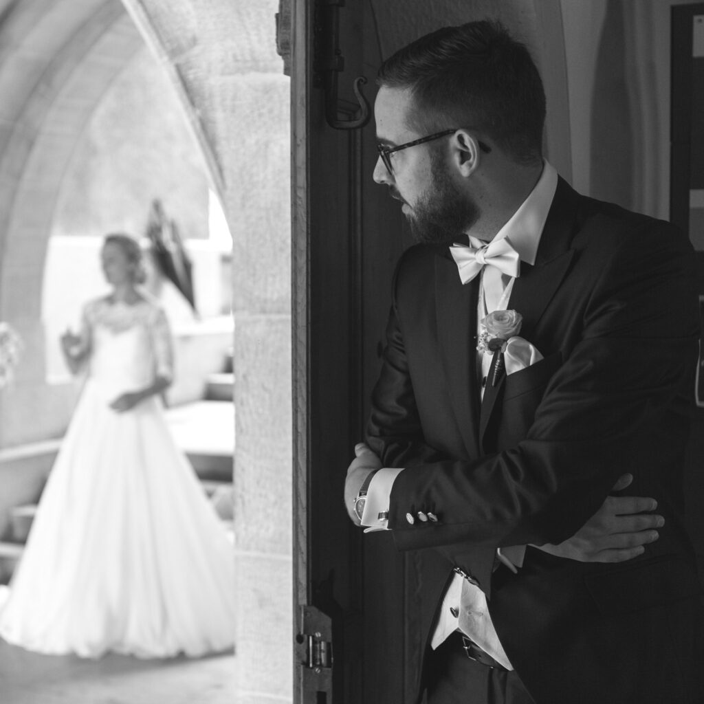 A black and white photo of a groom peering around a corner at his bride