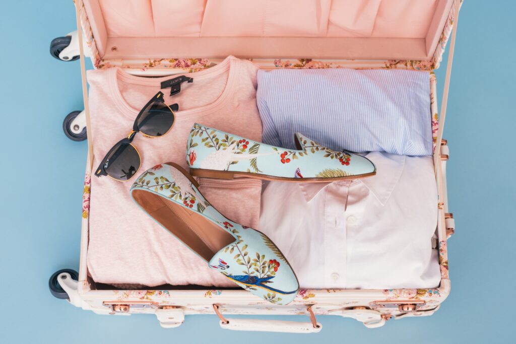 top down look at pink suitcase with pastel shoes, sunglasses, and clothing. 